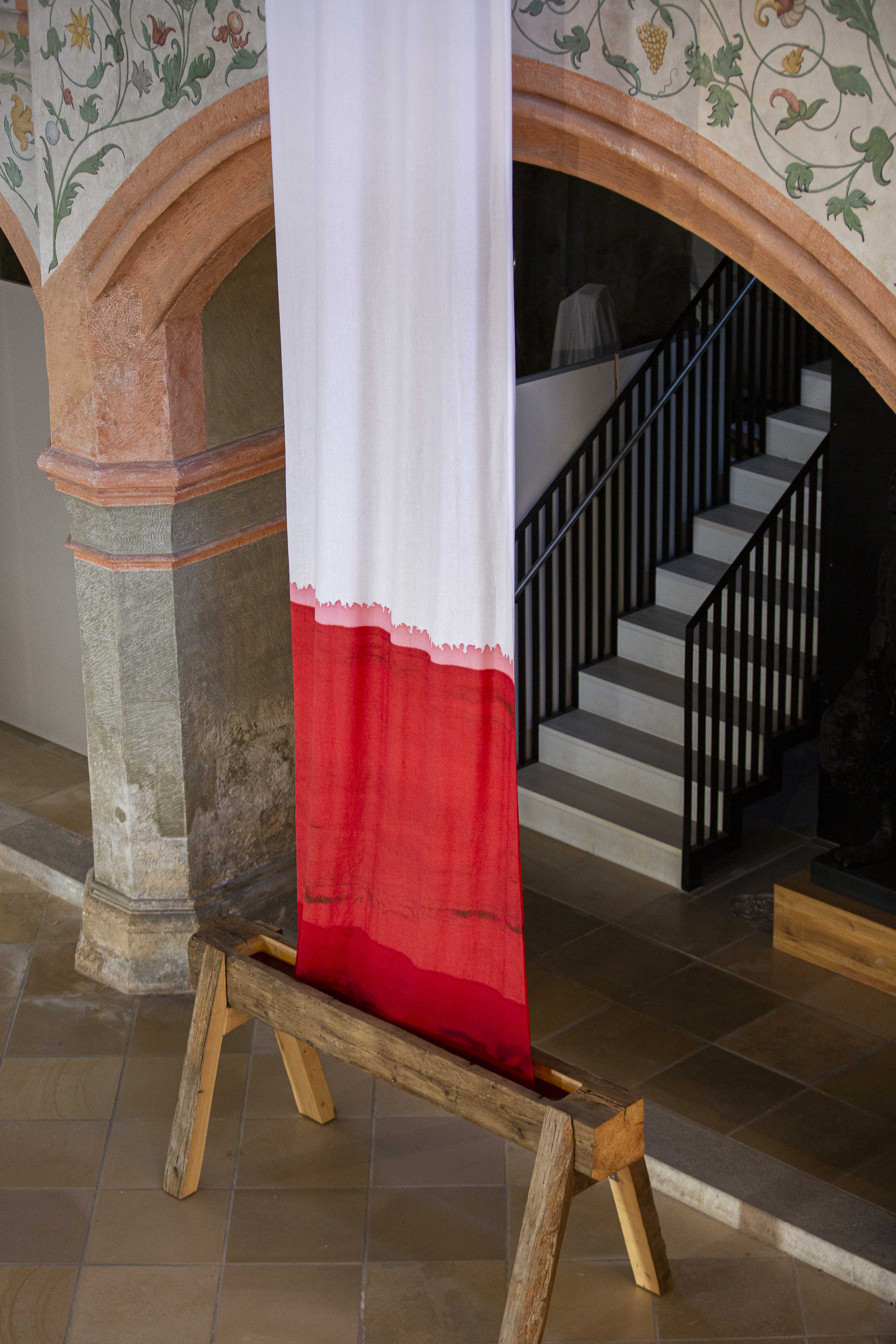 Langes, weiß-rotes Stoffband hängt in der Museumskirche St. Ulrich vor einem gemauerten Bogen und einer Treppe mit schwarzem Geländer.